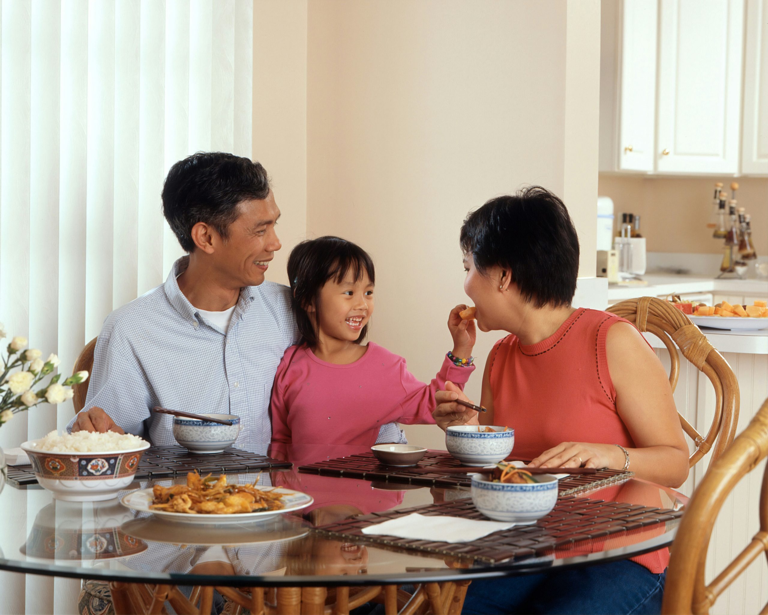 An Asian family, an adult male and female are seated around a table eating a meal with a young female standing in between the adults. Photographer Rhoda Baer