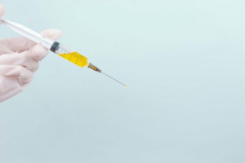 the-doctors-hand-in-a-white-medical-glove-holds-a-syringe-with-a-yellow-medicine-on-a-blue-background-the-medicine-place-for-an-inscription-vaccine-covid-19-coronavirus-stockpack-unsplash The doctor's hand in a white medical glove holds a syringe with a yellow medicine on a blue background. The medicine. Place for an inscription. Vaccine. Covid 19. Coronavirus.