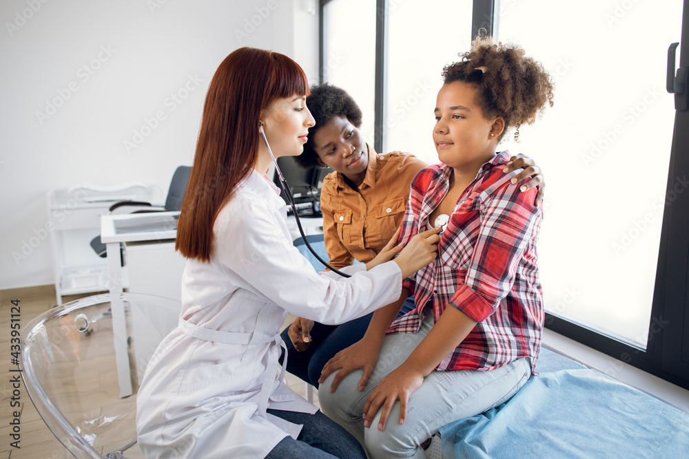 Flu, cough, asthma concept. Female pediatrician doctor listens to lungs of African-American 12-aged girl with stethoscope. Sick teen mixed race girl with asthma, supporting by her worried mom
